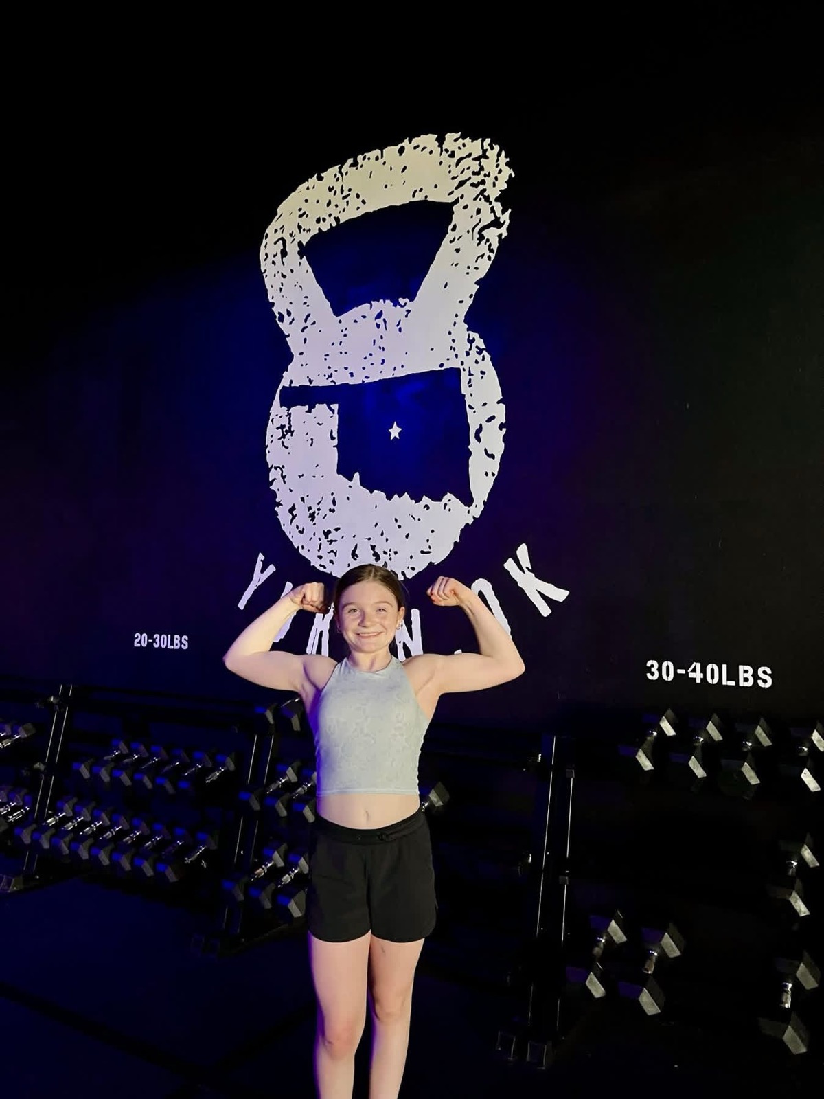 A girl flexes in front of the kettlebell wall and Yukon, OK mural at Arize Training Club.