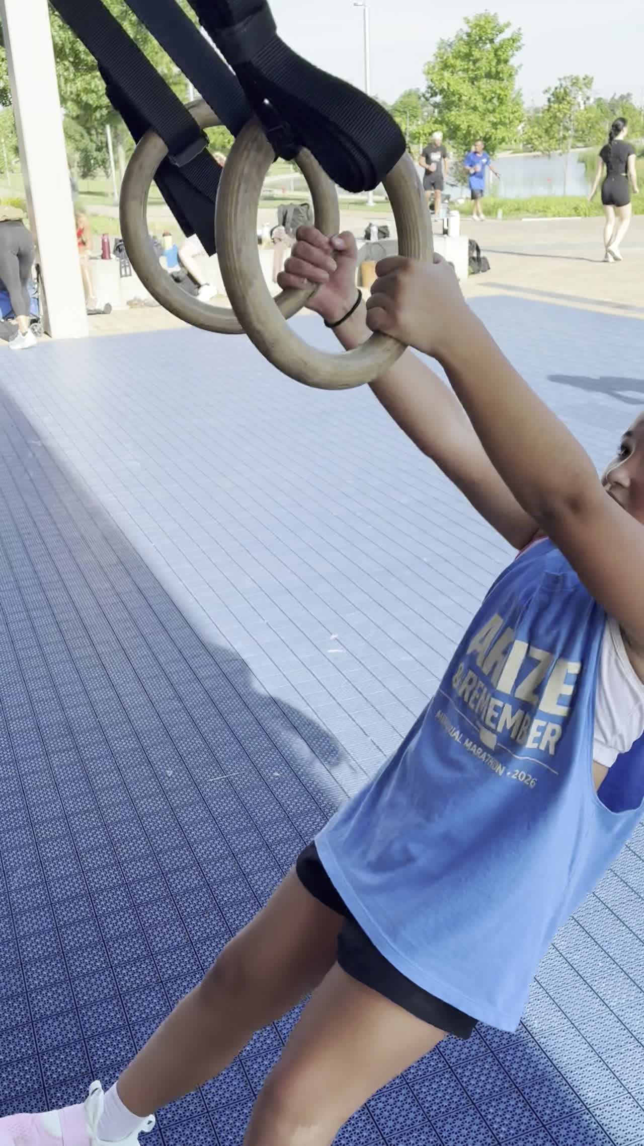 A kid on gymnastic rings wearing an Arize tank, outdoors.